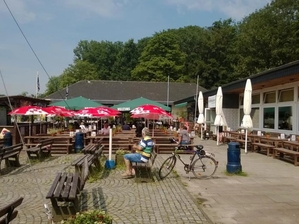 Se(h)-Restaurant Unterbacher See Sonnige Terrasse eines Restaurants am See mit rot-weißen Sonnenschirmen, Holzsitzbänken und Fahrrädern.
