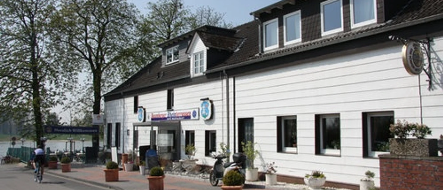 Baumberger Rheinterrassen in Monheim am Rhein Restaurant mit weißer Fassade, Blumenkübeln und angrenzender Terrasse mit Aussicht auf den Rhein.