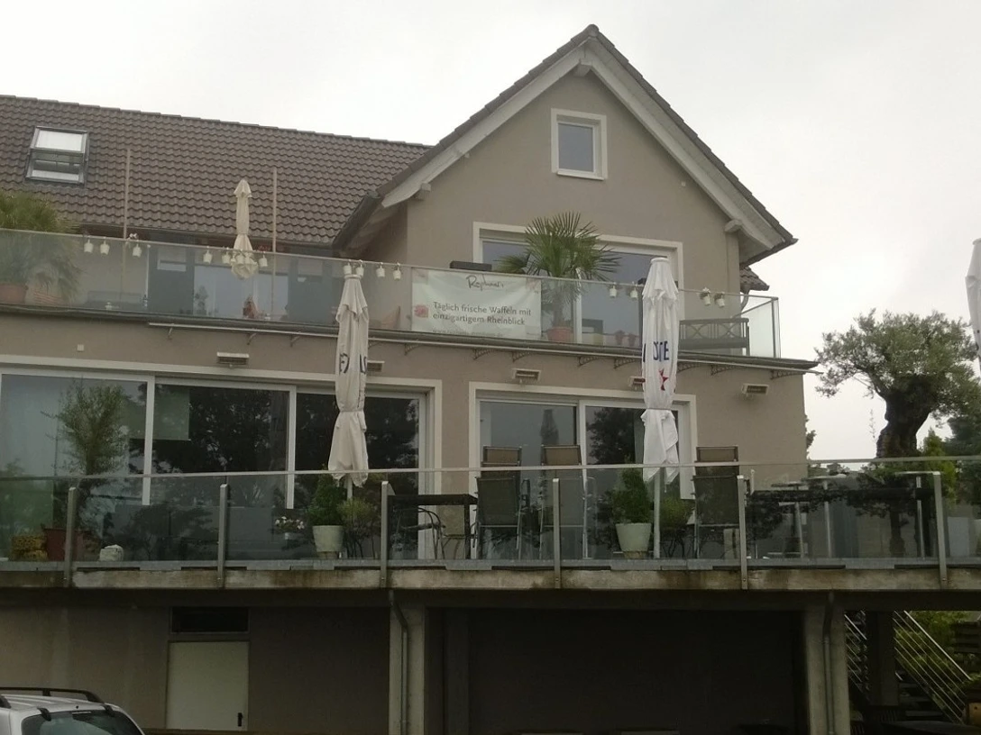 Zweistöckiges Gebäude mit großer Terrasse und zahlreichen Schirmen, umgeben von Pflanzen und Bäumen.