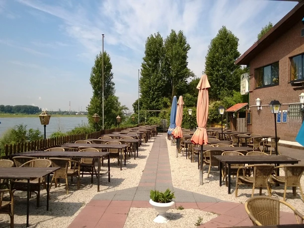 Ein Restaurant mit Außenterrasse und Blick auf einen Fluss bietet Sitzplätze zwischen bunten Sonnenschirmen.