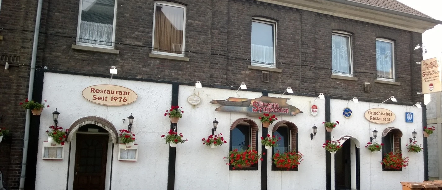 Taverna Symposion Das Bild zeigt ein traditionelles Restaurantgebäude mit weißen Wänden und dekorativen Blumen.
