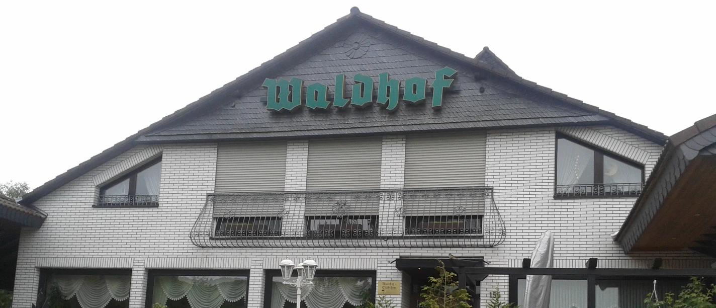 Café Restaurant Waldhof Weißes Backsteingebäude mit grünem Waldhof-Schild, Fenster mit Vorhängen und Laterne im Vordergrund.