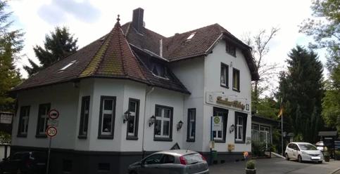 Landhaus Stolberg Ein zweistöckiges Landhaus mit Satteldach und Gaube, umgeben von Bäumen und mit parkenden Autos davor.