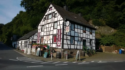 Fachwerkhaus mit Gaststätte, von Wald umgeben, bei strahlendem Sonnenschein an einer Straßenecke.