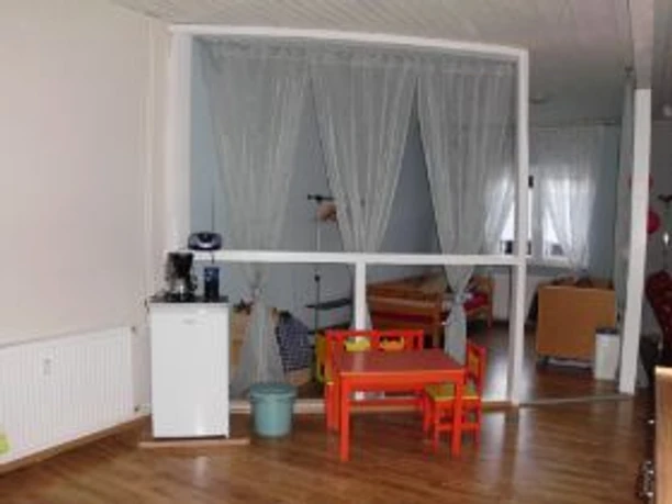 Kinderbereich mit kleinem Tisch und bunten Stühlen in einem hellen Raum mit Vorhangtrennung.