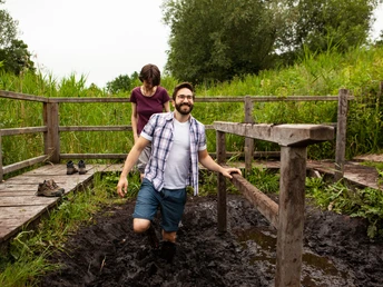 Zwei Menschen nutzen ein hölzernes Moortretbecken in einer grünen, von Bäumen umgebenen Umgebung.
