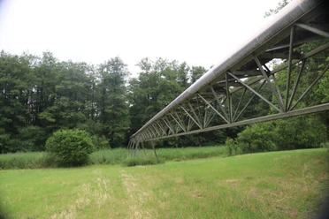 Moorpipeline Eine hohe Metallstruktur, die durch eine grüne Wiesenlandschaft und vor dichter Waldkulisse führt.
