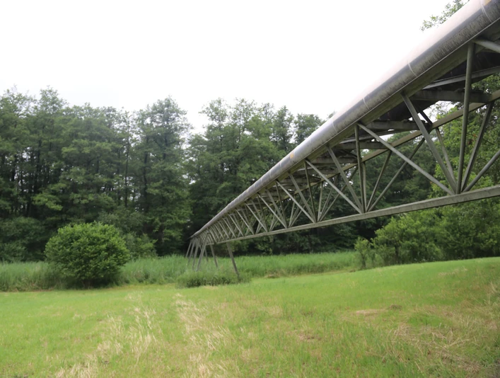 Moorpipeline Eine hohe Metallstruktur, die durch eine grüne Wiesenlandschaft und vor dichter Waldkulisse führt.