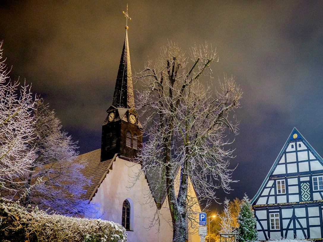 St. Stephan Winter Die winterliche Kirche St. Stephan in verschneiter, abendlicher Landschaft, flankiert vom Fachwerkhaus.