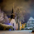 St. Stephan Winter Die winterliche Kirche St. Stephan in verschneiter, abendlicher Landschaft, flankiert vom Fachwerkhaus.