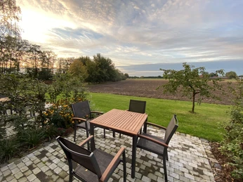 Terrasse mit Holztisch und Stühlen; dahinter grüne Wiese und gepflügtes Feld bei Sonnenuntergang.
