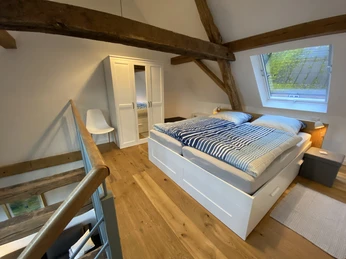 Dachzimmer mit rustikalen Holzbalken, Doppelbett, Kleiderschrank, Fenster mit Ausblick auf grüne Natur.