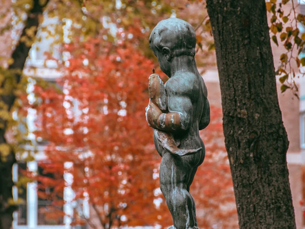 Fischpüddelchen im Herbst Bronzestatue eines Kindes, Herbstblätter im Hintergrund, steht auf einem Steinpodest.