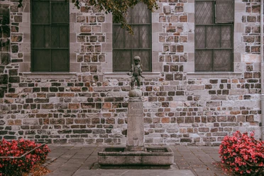 Ein kleiner Junge aus Stein, das „Fischpüddelchen“, steht auf einem Brunnen vor einer historischen Gebäudefassade, umgeben von roten Blumen.