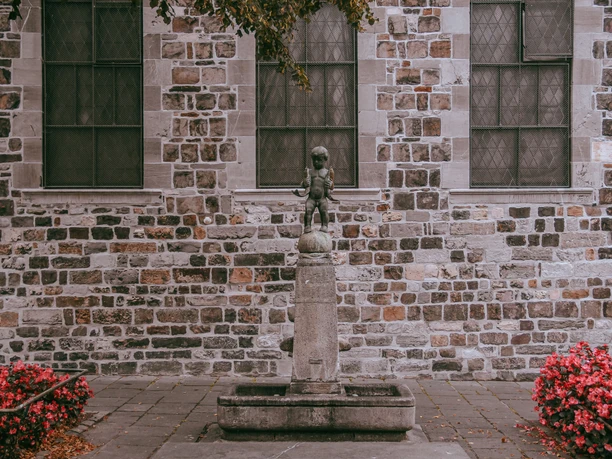 Ein kleiner Junge aus Stein, das „Fischpüddelchen“, steht auf einem Brunnen vor einer historischen Gebäudefassade, umgeben von roten Blumen.