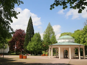 Brunnentempel mit Musikmuschel inmitten eines gepflegten Parks mit grünen Bäumen und blauen Himmel.