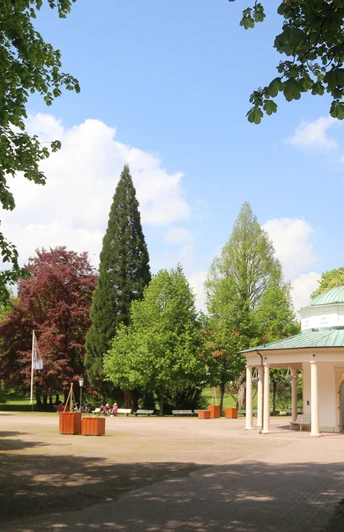 Brunnentempel mit Musikmuschel Ein historischer Pavillon mit grüner Kuppel steht in einem parkähnlichen Umfeld voller Bäume.