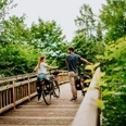 Zwei Personen schieben Fahrräder über einen hölzernen Steg, umgeben von üppigem Grün und Bäumen.