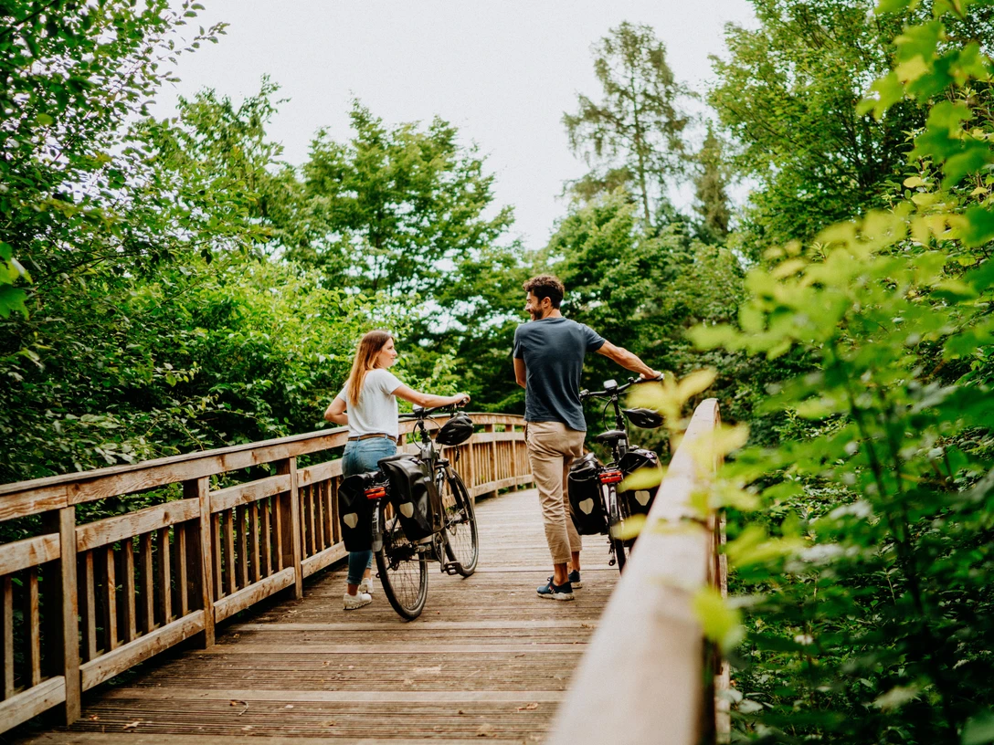 Brakel - Rheder Zwei Personen schieben Fahrräder über einen hölzernen Steg, umgeben von üppigem Grün und Bäumen.