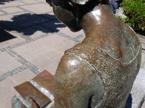Blick von hinten über die Schulter einer lesenden Bronze-Statue.