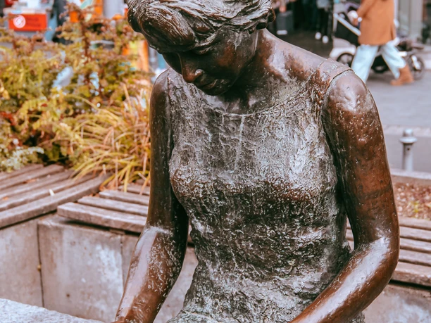 Nahaufnahme von einer lesenden Brone-Statue mit Blumen, einer Straße und der Buchhandlung im Hintergrund.