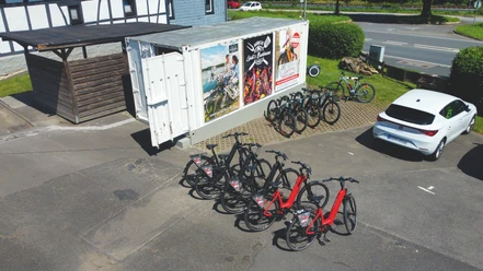Mehrere Fahrräder, teils rot, stehen in einem Abstellbereich neben einem Container und einem Pkw.