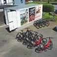 Lüdenbach_E-BIke-Verleih Mehrere Fahrräder, teils rot, stehen in einem Abstellbereich neben einem Container und einem Pkw.