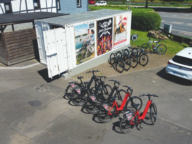 Lüdenbach_E-BIke-Verleih Mehrere Fahrräder, teils rot, stehen in einem Abstellbereich neben einem Container und einem Pkw.