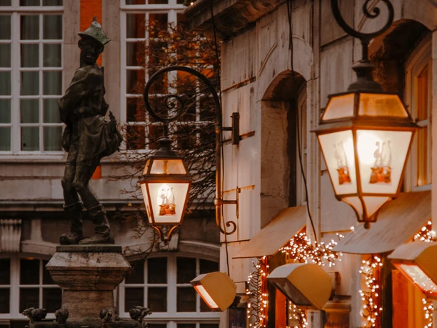 Hühnerdieb Lichterglanz Leuchtende Straßenlaternen an einer historischen Gebäudewand, die mit Lichterketten geschmückt sind. Vor der Wand steht die Statue eines Jungen mit Huhn im Arm.
