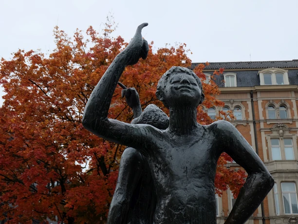 Klenkes-Denkmal Herbst Bronze-Figuren mit erhobenem kleinen Finger vor einem herbstlichen Baum und einem Gebäude.