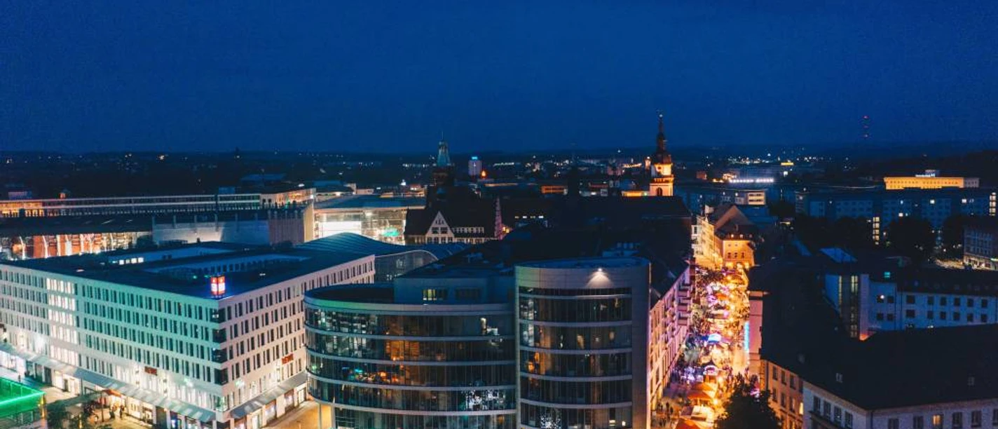 brauereimarkt_07466-ernestouhlmann.jpg Abendliche Luftaufnahme von Linz mit beleuchteten, modernen Gebäuden und lebhaftem Straßenleben.