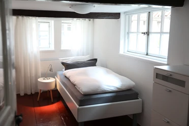 Helles Einzelzimmer mit weißem Bett, Holzbalken an der Decke, Fenster und einem Nachttisch mit Lampe.