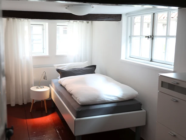 Helles Einzelzimmer mit weißem Bett, Holzbalken an der Decke, Fenster und einem Nachttisch mit Lampe.