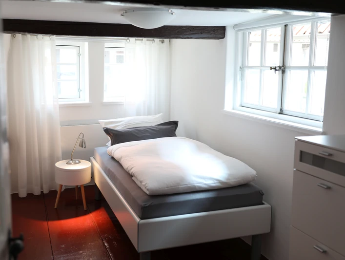 Korona_3.jpg Helles Einzelzimmer mit weißem Bett, Holzbalken an der Decke, Fenster und einem Nachttisch mit Lampe.