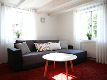 Helles Wohnzimmer mit grauem Sofa, rotem Teppich und rundem weißen Tisch unter Holzdeckenbalken.