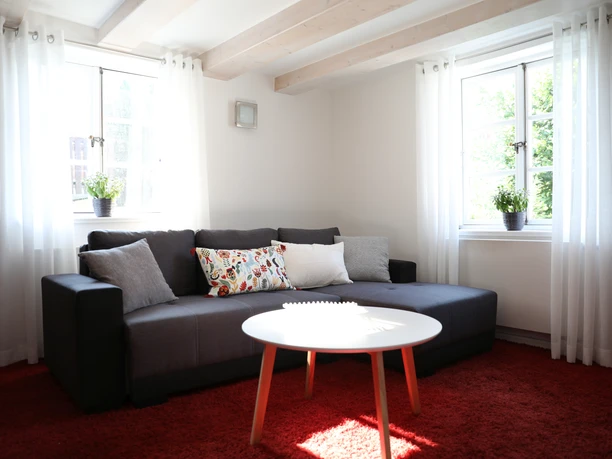 Helles Wohnzimmer mit grauem Sofa, rotem Teppich und rundem weißen Tisch unter Holzdeckenbalken.