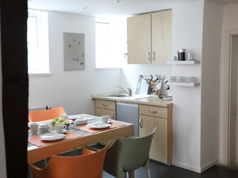 Korona_4.jpg Helle, modern eingerichtete Küche mit Esstisch und buntem Geschirr vor zwei Fenstern.