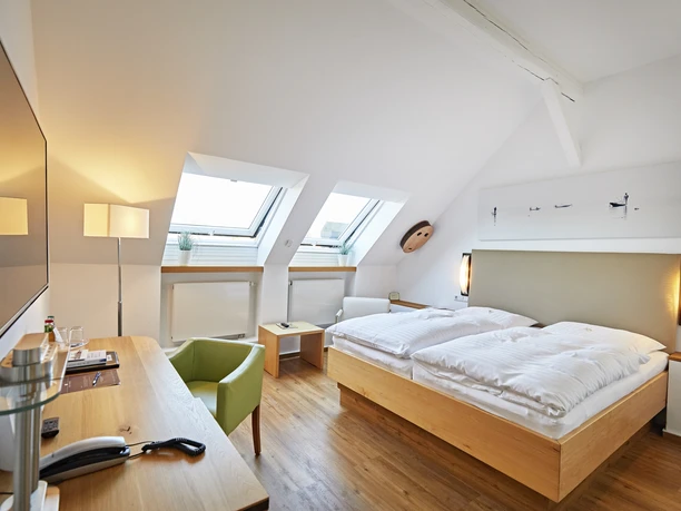 Modern eingerichtetes Hotelzimmer mit zwei Dachfenstern, Doppelbett, Schreibtisch und grünem Sessel.