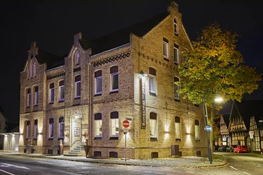 Historisches Backsteingebäude bei Nacht, stimmungsvoll beleuchtet, mit Bäumen und Fachwerk im Hintergrund.