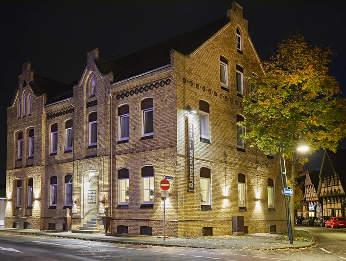 Wartburg_5.jpeg Historisches Backsteingebäude bei Nacht, stimmungsvoll beleuchtet, mit Bäumen und Fachwerk im Hintergrund.