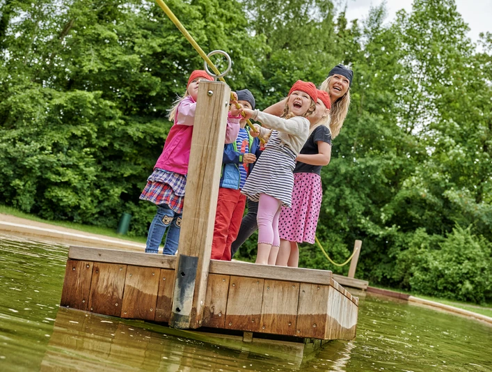 Spielpark Wingst Piratenwelt Kinder mit Piratenbändern überqueren lachend das Wasser auf einem Floß im Spielpark Wingst.Children with pirate ribbons laugh as they cross the water on a raft in the Wingst play park.Børn med piratbånd griner, mens de krydser vandet på en tømmerflåde i Wingst legepark.Kinderen met piratenlintjes lachen terwijl ze het water oversteken op een vlot in het speelpark Wingst.