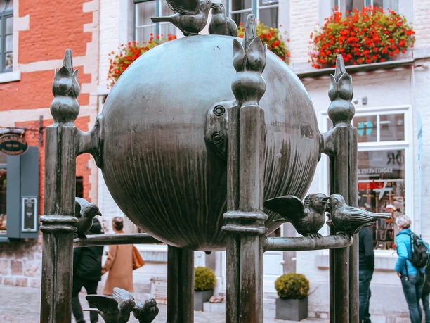 Möschebrunnen im Herbst Brunnen, der aus einer Bronze-Kugel mit sitzenden Vögeln und mehreren Bronze-Säulen mit weiteren Vogel-Figuren besteht. Im Hintergrund steht ein Haus mit historischer Fassade und roten Blumen.