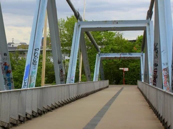 Heuschreckenbrücke Eine moderne Brücke mit grauen, mit Graffiti bedeckten Stahlträgern überquert einen Gehweg, mit dem berühmten Kölner Dom, der zwischen Bäumen im Hintergrund aufragt.A modern bridge with gray, graffiti-covered steel girders crosses a sidewalk, with the famous Cologne Cathedral towering between trees in the background.