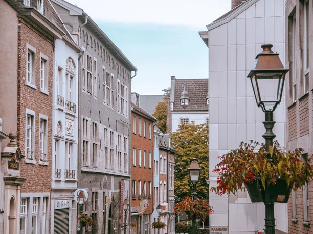 Gepflasterte Gasse zwischen bunten Häuserreihen und historischen Straßenlaternen, die mit Blumenkästen geschmückt sind.