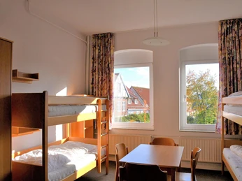 Kloster_Wiedenbrück_1.jpg Helles Hostelzimmer mit zwei Etagenbetten, Tisch und Stühlen, Blick auf Bäume und Nachbargebäude.