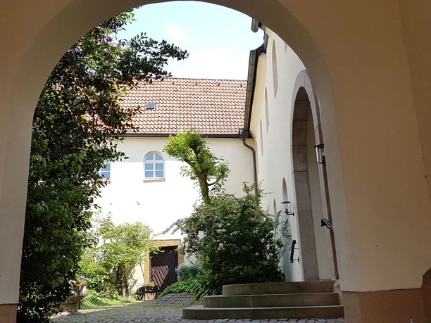 Durch einen Torbogen blickt man auf einen Innenhof mit blühenden Pflanzen und einem historischen Gebäude.