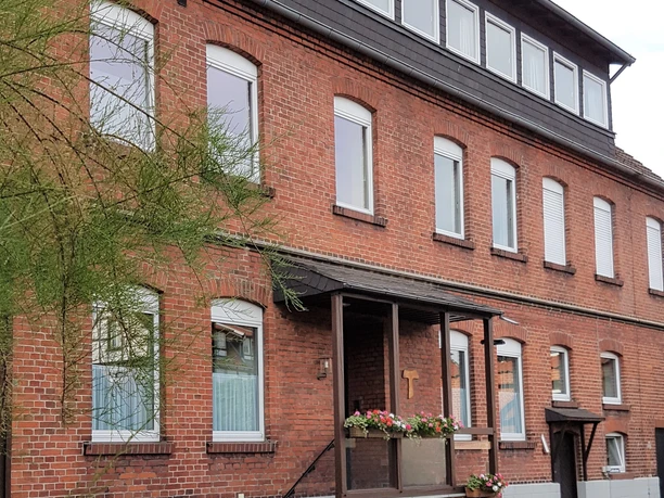Ein dreistöckiges Backsteingebäude mit weißen Fensterrahmen und dekorativem Blumenschmuck am Eingang.