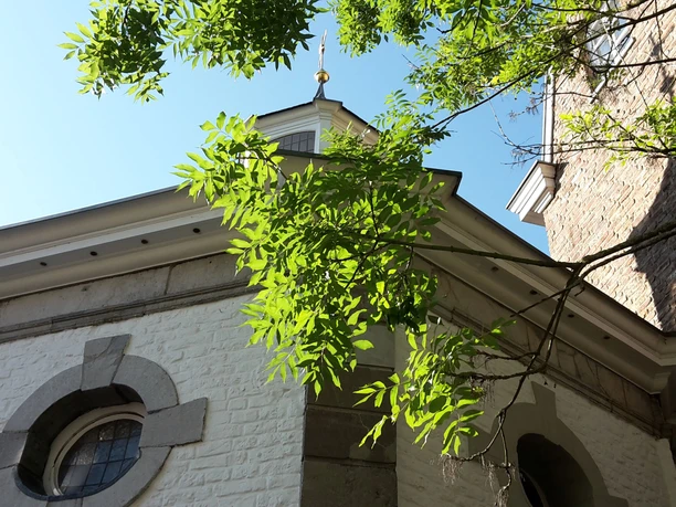 Achteckige, weiße Kapelle hinter einem grünen Baum unter blauem Himmel.