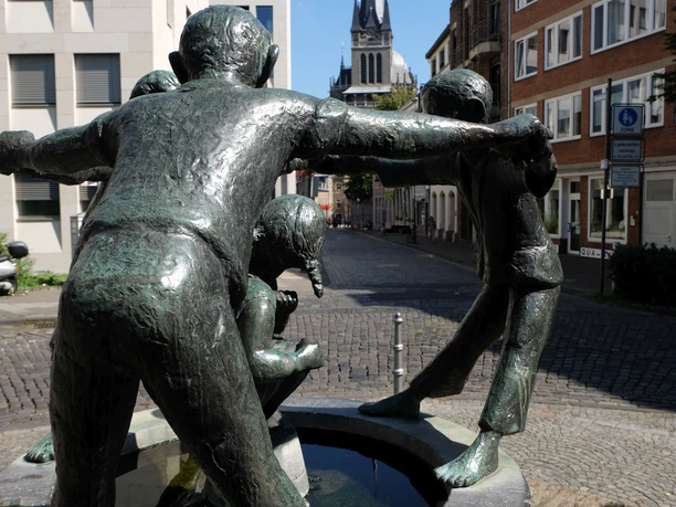Eine Skulptur zeigt drei bronzene, lachende Figuren, die sich an den Händen halten und ein sitzendes Kind umgeben. Im Hintergrund befinden sich Wohnhäuser, eine Gasse sowie die Turmspitze des Aachener Doms.