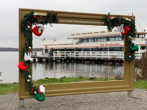winter-wunder-welt-2021-bilderrahmen.jpg Weihnachtlich geschmückter Bilderrahmen mit Blick auf das Fahrgastschiff Bad Zwischenahn am See.